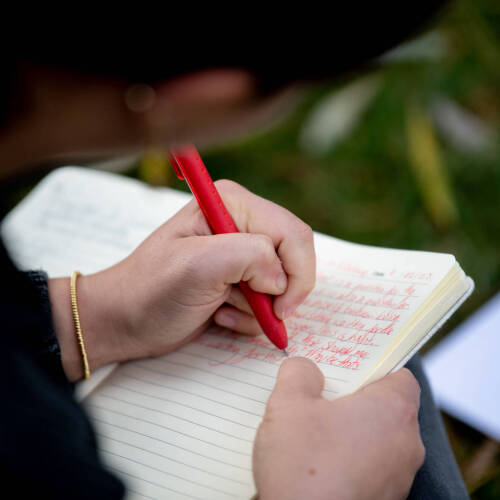 Student writing in notebook with red pen